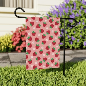 May include: A pink garden flag featuring a repeating pattern of red strawberries with green stems and white flowers. The flag is displayed on a black metal stand in a grassy yard, with flowering bushes in the background.