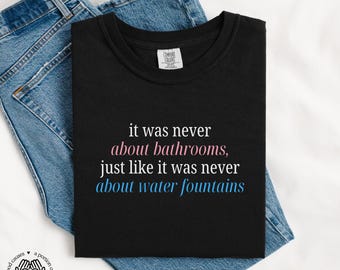May include: Black t-shirt with white text that reads "it was never about bathrooms, just like it was never about water fountains".