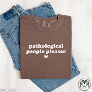 May include: A brown t-shirt with the text "pathological people pleaser" in white letters and a small white heart.