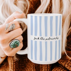 May include: A white ceramic mug featuring vertical light blue stripes and the black text "fuck the patriarchy" printed in the center. The mug has a curved handle on the left side and a smooth, cylindrical body with a clean, minimalist design.