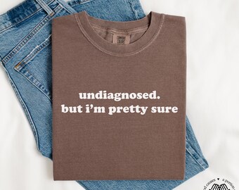May include: A brown t-shirt with the text "undiagnosed. but i'm pretty sure" printed in white.