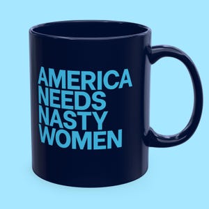 May include: A dark navy blue ceramic mug featuring the text AMERICA NEEDS NASTY WOMEN printed in a bold, light blue sans-serif font. The mug has a smooth, glossy finish, a cylindrical body, and a curved handle on the right side.