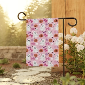 May include: A rectangular garden flag featuring a repeating pattern on a light pink background. The design includes white ghosts, orange pumpkins, pink bows, purple spider webs, and small white skulls arranged in a grid-like layout.