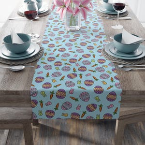 May include: A light blue Easter-themed table runner with a repeating pattern of decorated eggs, carrots, and bunny silhouettes. The runner is placed on a wooden table set for a meal, with plates, bowls, and wine glasses.