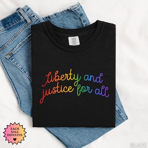 May include: Black t-shirt with the rainbow text "liberty and justice for all." The shirt is folded on top of blue denim jeans. A small tag on the shirt reads "Comfort Colors." A pink and orange starburst graphic is in the lower left corner.