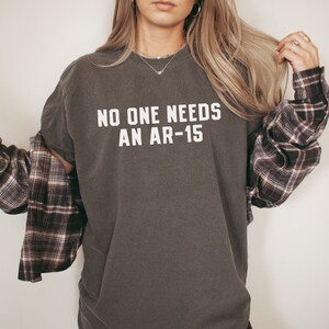 May include: Dark gray t-shirt with the text "NO ONE NEEDS AN AR-15" in white block letters. The shirt is paired with a brown and burgundy plaid flannel shirt. The image is a medium shot.