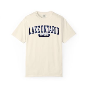 May include: A cream-colored short-sleeve t-shirt featuring the dark blue text LAKE ONTARIO in an arched collegiate font, with the text EST 1660 centered below inside a dark blue rounded rectangular border on the chest of the garment.