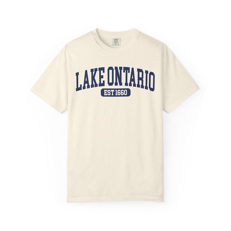 May include: A cream-colored short-sleeve t-shirt featuring the dark blue text LAKE ONTARIO in an arched collegiate font, with the text EST 1660 centered below inside a dark blue rounded rectangular border on the chest of the garment.