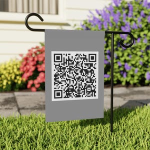 May include: A gray garden flag with a large black and white QR code. The flag is displayed on a black metal stand in a grassy yard, with colorful flowers in the background.