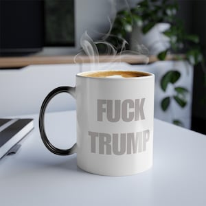 May include: White ceramic mug with the text "FUCK TRUMP" in gray. The mug has a black handle and is filled with a hot beverage, with steam rising from the top. The mug is on a white surface.