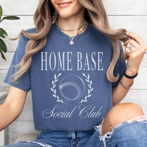 May include: A blue t-shirt with a white graphic of a baseball with a wreath around it and the text "Home Base Social Club".