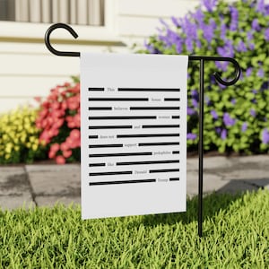May include: A white garden flag with black text that reads "This house believes women and does not support pedophiles like Donald Trump." The flag is hanging on a black metal stand in a garden setting with flowers.