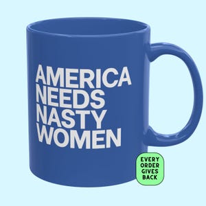 May include: A blue ceramic mug featuring the white text AMERICA NEEDS NASTY WOMEN printed in a bold sans-serif font. A small light green rounded square badge in the bottom right corner displays the black text EVERY ORDER GIVES BACK.
