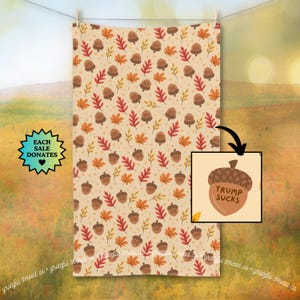 May include: A beige towel with a repeating pattern of acorns and fall leaves in shades of brown, orange, and red. The acorns have the text "TRUMP SUCKS" on them. The towel is hanging on a clothesline.