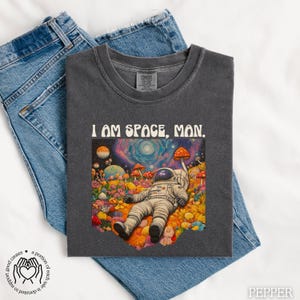 May include: A dark gray t-shirt with a colorful psychedelic graphic of an astronaut in a field of mushrooms. The text "I AM SPACE, MAN." is printed in white on the shirt.