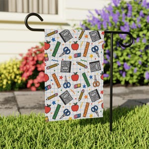 May include: A rectangular garden flag hanging on a black metal stand, featuring a white grid pattern background with a repeating school supply print including red apples, yellow pencils, blue scissors, green rulers, pink erasers, and black notebooks.
