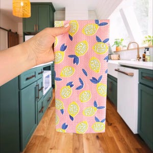 May include: Pink kitchen towel with a pattern of yellow lemons, blue leaves, and the text "FUCK TRUMP" in yellow. The towel is held up in front of a kitchen setting with green cabinets and wooden floors.
