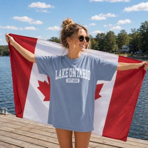 May include: A light blue short-sleeve t-shirt featuring the white text "LAKE ONTARIO" and "EST 1660" printed on the chest. The shirt is held against a large Canadian flag with red and white vertical stripes and a central red maple leaf design.