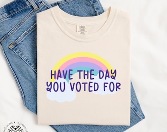 May include: A beige t-shirt with a rainbow and the text "Have the day you voted for".