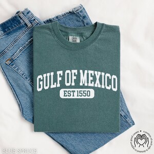 Puede incluir: Una camiseta azul turquesa con texto blanco que dice "GULF OF MEXICO EST 1550".