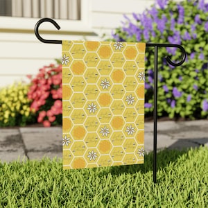 May include: A yellow garden flag with a honeycomb pattern, white flowers, and the text "Fuck Trump". The flag is hanging on a black metal stand in a garden setting with green grass and colorful flowers.