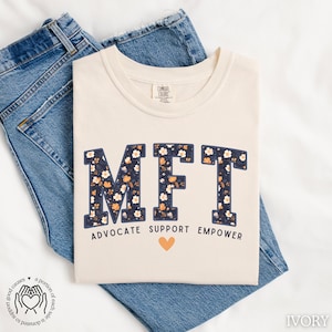 May include: A beige t-shirt with a floral print of the letters "MFT" and the words "Advocate Support Empower" below. A small heart is between the words "Support" and "Empower".
