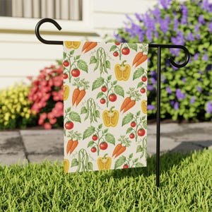 May include: A rectangular garden flag hanging on a black metal stand, featuring a repeating pattern of red tomatoes, orange carrots, yellow bell peppers, and green bean pods on a cream background with the word trump written in cursive script.