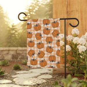 May include: A garden flag featuring a repeating pattern of orange pumpkins with green stems on a white and orange checkered background. The flag is displayed on a black metal stand in an outdoor setting, with greenery and flowers in the background.