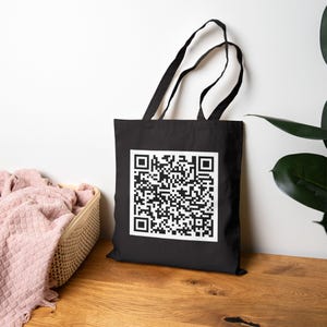 May include: Black tote bag with a large white square containing a QR code. The bag has black handles and is sitting on a wooden surface. A basket with a pink blanket is to the left of the bag.