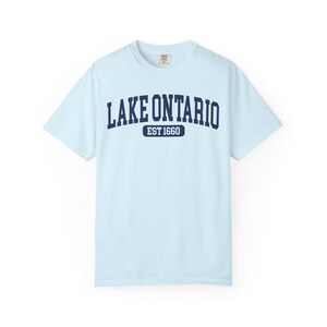 May include: A light blue short-sleeve t-shirt featuring the dark blue text LAKE ONTARIO in an arched font, with the text EST 1660 inside a dark blue rounded rectangle below it. The shirt has a crew neckline and a standard fit design.