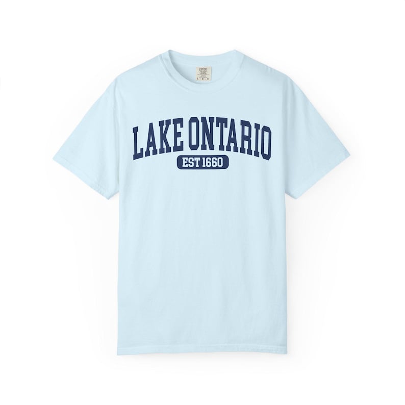 May include: A light blue short-sleeve t-shirt featuring the dark blue text LAKE ONTARIO in an arched font, with the text EST 1660 inside a dark blue rounded rectangle below it. The shirt has a crew neckline and a standard fit design.