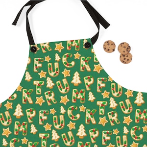 May include: Green apron with a repeating pattern of gingerbread cookie letters spelling out a phrase, along with star and Christmas tree cookies. The apron has black straps and buttons. Three chocolate chip cookies are in the upper right corner.
