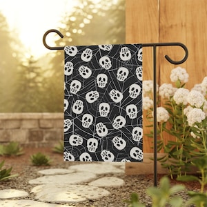 May include: A rectangular garden flag featuring a repeating pattern of white skulls and spiderwebs on a black background. Each skull has the letters FDT printed in black on the forehead. The flag hangs from a black metal garden flag stand.