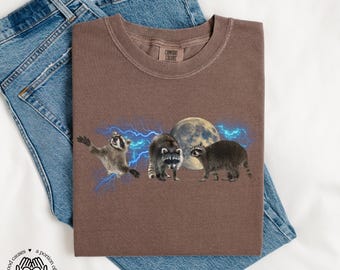May include: A brown t-shirt with a graphic of three raccoons in front of a full moon and lightning bolts. The text "COMFORT COLORS" is on the shirt.