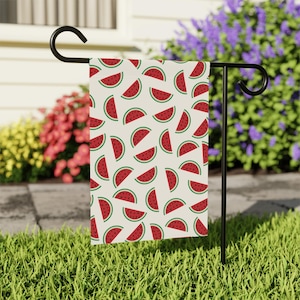May include: A rectangular garden flag with a repeating pattern of red watermelon slices on a cream background. The flag is displayed on a black metal stand in a grassy yard, with flowers and a house in the background.