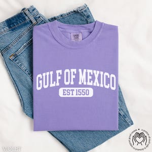 Puede incluir: Una camiseta morada con texto blanco que dice "GULF OF MEXICO EST 1550". La camiseta est&aacute; puesta sobre una superficie blanca con un par de pantalones vaqueros azules.