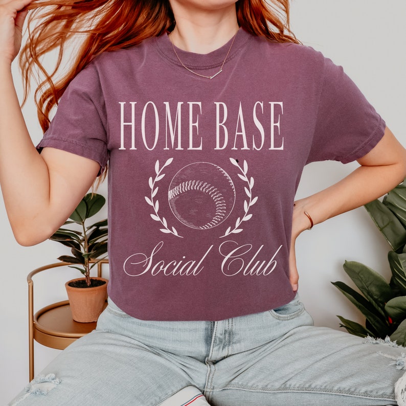 May include: A purple t-shirt with the text "HOME BASE Social Club" and a white baseball graphic with a laurel wreath.