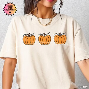 May include: Ivory-colored t-shirt featuring three orange pumpkin illustrations with the text "fuck trump" above each pumpkin. The shirt has short sleeves and a round neckline. A small tag with the text "EACH SALE DONATES" is visible.