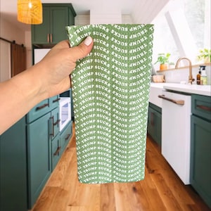 May include: Green kitchen towel with the words "TRUMP SUCKS" repeated in a pattern. The towel is held in front of a kitchen with green cabinets and a wooden floor.