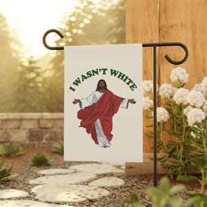 May include: A garden flag featuring an illustration of Jesus with outstretched arms, wearing a white robe and a red cloak. The text "I WASN'T WHITE" is printed above the image in green letters. The flag is displayed on a black metal stand.