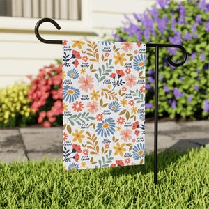 May include: A rectangular garden flag hanging on a black metal stand, featuring a white background with a repeating floral pattern in shades of red, yellow, and blue, interspersed with the repeated black text "FUCK TRUMP" throughout the design.
