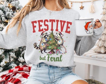 May include: A light gray sweatshirt with the text "FESTIVE" in red block letters above a graphic of two raccoons and a Christmas tree. The text "but Feral" is below the graphic in green. A white mug with a Santa Claus graphic is held in the hand.