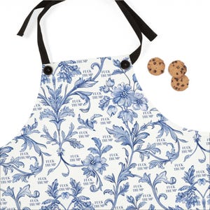 May include: White apron with a blue floral design and the text "FUCK TRUMP" repeated throughout. The apron has black ties and buttons. Three chocolate chip cookies are next to the apron.