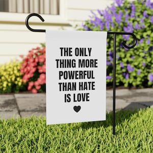 May include: A white garden flag with the text "THE ONLY THING MORE POWERFUL THAN HATE IS LOVE" in black, with a black heart symbol. The flag is displayed on a black metal stand in a garden setting with colorful flowers.