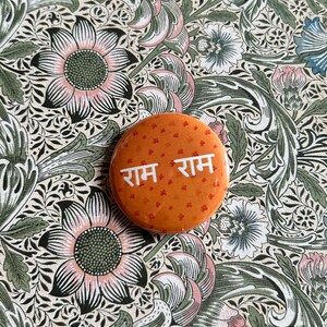 May include: A round orange button with the words "Ram Ram" in white lettering. The button is decorated with small red symbols.