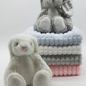 May include: A stack of four plush blankets in blue, white, grey, and pink. A white plush bunny sits in front of the stack. A grey plush bunny with the text "Baby Booth" embroidered on its ear sits on top of the stack.