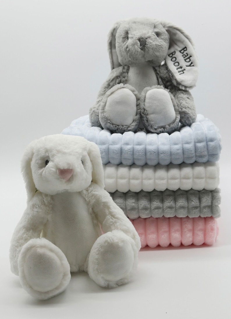 May include: A stack of four plush blankets in blue, white, grey, and pink. A white plush bunny sits in front of the stack. A grey plush bunny with the text "Baby Booth" embroidered on its ear sits on top of the stack.