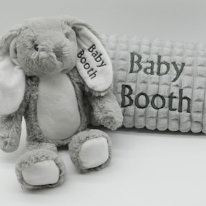 May include: A grey plush bunny with white ears and a grey and white textured blanket. The blanket and bunny are both embroidered with the name "Baby Booth".