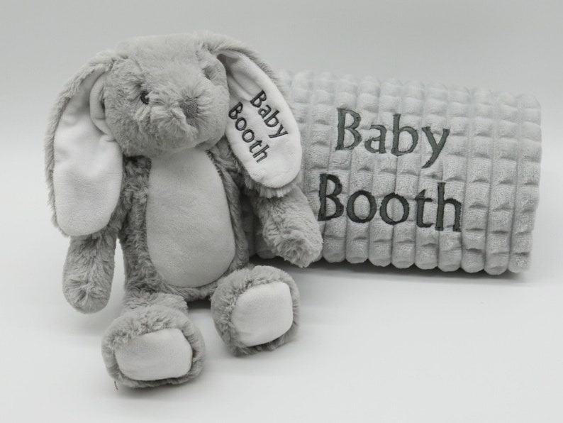 May include: A grey plush bunny with white ears and a grey and white textured blanket. The blanket and bunny are both embroidered with the name "Baby Booth".