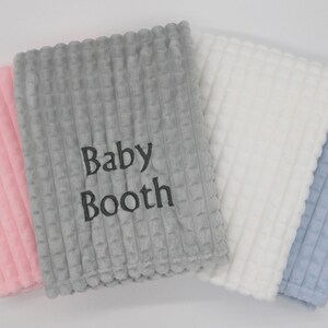 May include: Four baby blankets in different colours: pink, grey, white and blue. The grey blanket has the text "Baby Booth" embroidered on it.